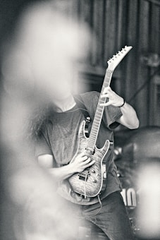 A person playing an electric guitar with a detailed wood finish, partially obscured by what appears to be smoke or a blurred element in the foreground. The image has a high contrast and a monochromatic tone, emphasizing textures and shadows.