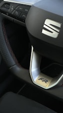 Close-up view of a car steering wheel displaying a prominent logo in the center. The steering wheel features a sporty design with a silver trim and is accompanied by buttons for various controls. The stitching on the wheel is visible, indicating a detailed and refined finish.