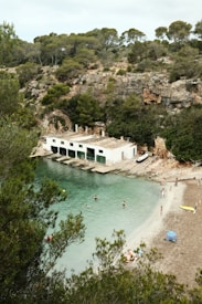 A serene cove with clear turquoise water and a sandy beach is surrounded by rocky cliffs and dense green pine trees. People are swimming and lounging on the beach, and a white building with green doors sits along the water's edge.