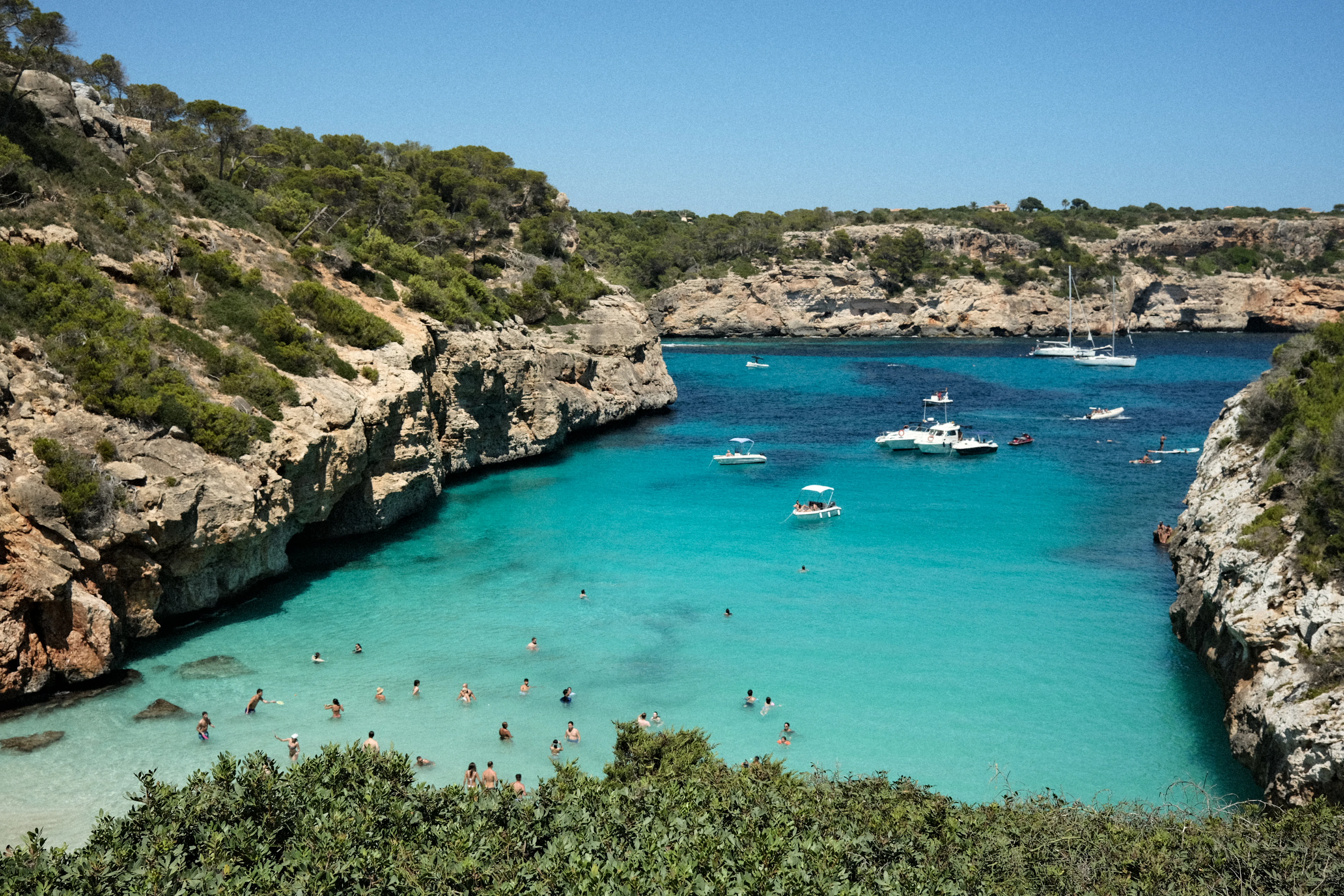Swimmers and anchored boats in a turquoise limestone cove ringed by pine-covered cliffs.