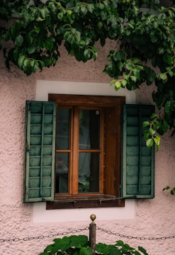 A rustic wooden window frame with open teal-colored shutters against a textured pale pink wall. Green foliage hangs above the window, with some leaves slightly covering the shutters. A brass finial and a black metal chain are positioned below the window.