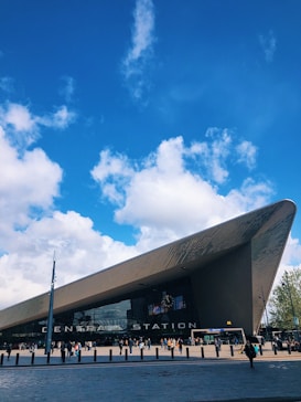 A modern and sleek architectural structure identified as a central station, with an angular roof, set against a vibrant blue sky scattered with white clouds. Numerous people can be seen approaching and entering the building, which is flanked by tall streetlamps and urban infrastructure.
