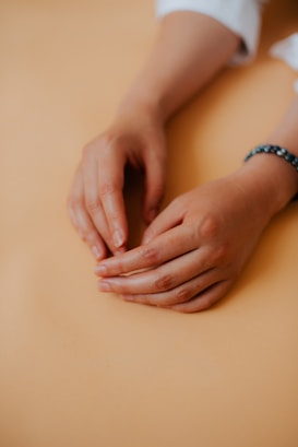 Hands are gently resting on a surface, with a bracelet visible on one wrist. The skin tones are warm and the light is soft, creating a serene ambiance.