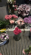 A display of an assortment of floral arrangements in various containers. The flowers include pink tulips, carnations, and purple hydrangeas. The flowers are arranged in metallic buckets and glass vases on tiers of white concrete blocks. There is a striped rug underneath the display, and some greenery adds contrast to the colorful flowers.