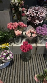 A display of an assortment of floral arrangements in various containers. The flowers include pink tulips, carnations, and purple hydrangeas. The flowers are arranged in metallic buckets and glass vases on tiers of white concrete blocks. There is a striped rug underneath the display, and some greenery adds contrast to the colorful flowers.