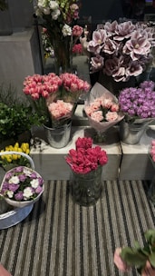 A display of an assortment of floral arrangements in various containers. The flowers include pink tulips, carnations, and purple hydrangeas. The flowers are arranged in metallic buckets and glass vases on tiers of white concrete blocks. There is a striped rug underneath the display, and some greenery adds contrast to the colorful flowers.