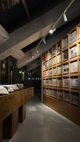 A cozy interior space featuring a wooden display of vinyl records arranged on a grid-like wooden shelf along one wall. The room is dimly lit with spotlights coming from the ceiling, illuminating the records and creating a warm, inviting atmosphere. A long wooden table runs parallel to the shelf, displaying additional records.