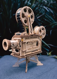 A vintage-style wooden model of a film projector with intricate details and engravings. The projector features large spools, a lens, and a stand. Set against a blurred, natural background.