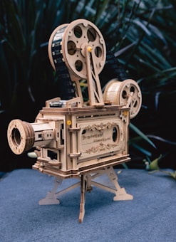 A vintage-style wooden model of a film projector with intricate details and engravings. The projector features large spools, a lens, and a stand. Set against a blurred, natural background.