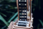 A close-up view of a film reel with a strip of film running through a wooden mechanism. The background is blurred, featuring green foliage.