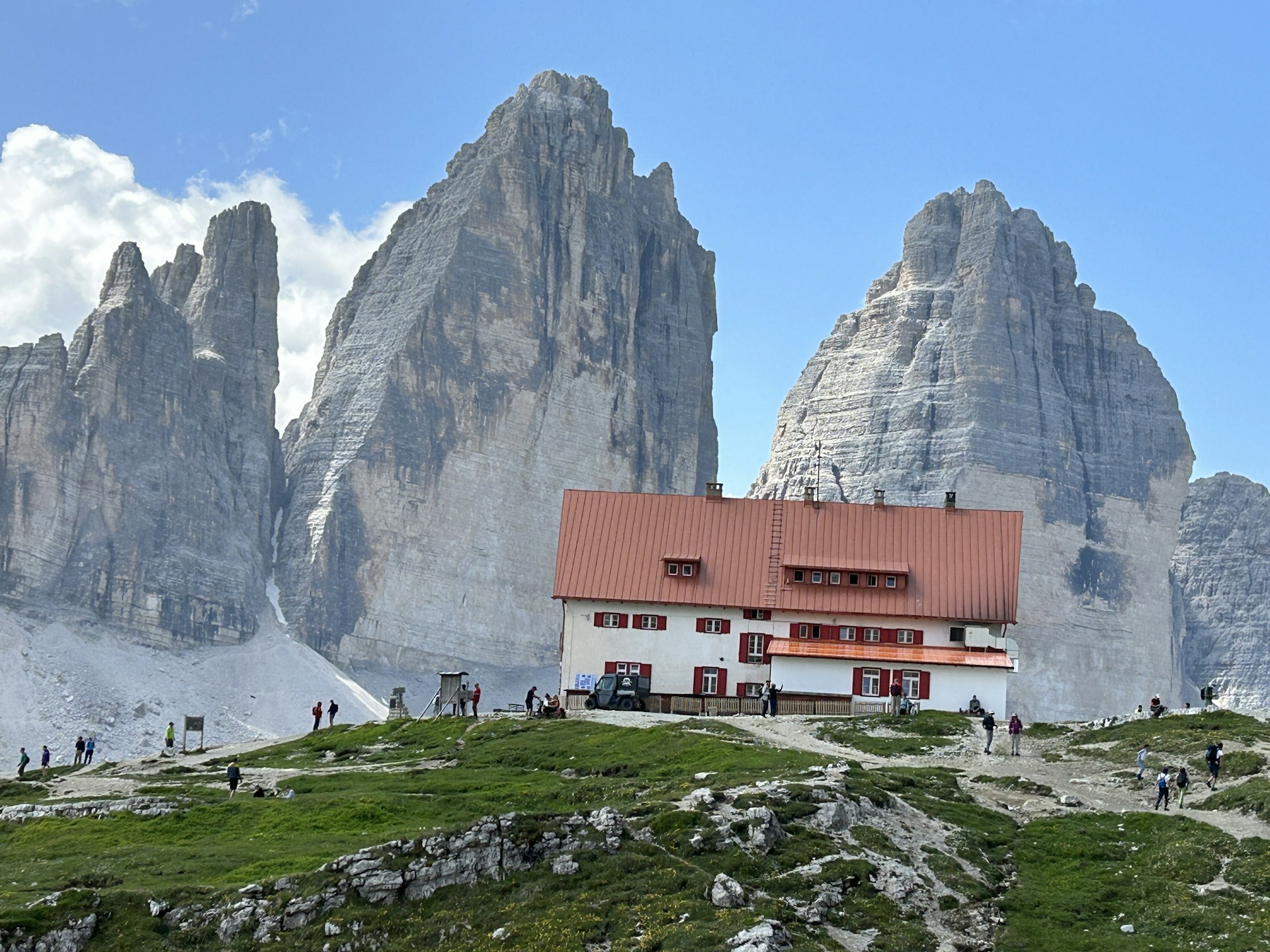 A mountain lodge with a red roof and white walls is situated at the base of towering rock formations under a clear blue sky. Several people are visible walking along a path leading to the lodge, surrounded by rugged terrain and patches of grass.