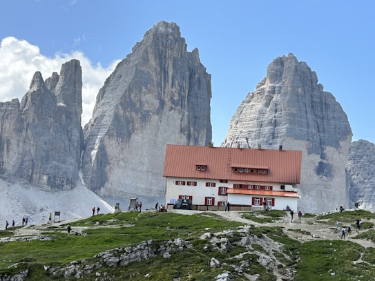 A mountain lodge with a red roof and white walls is situated at the base of towering rock formations under a clear blue sky. Several people are visible walking along a path leading to the lodge, surrounded by rugged terrain and patches of grass.