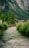 A serene river flows through a lush green valley, flanked by dense foliage and bushes. A rustic wooden house is nestled among the trees, with a backdrop of towering forested mountains.