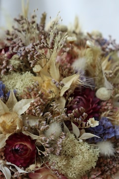 A collection of dried flowers and plants featuring a variety of textures and colors, including shades of red, purple, blue, beige, and yellow. The arrangement includes strawflowers, moss, and clusters of small blossoms, creating an intricate, natural composition.