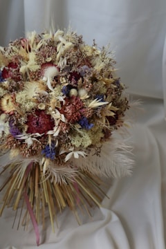 A bouquet of dried flowers, featuring a mix of textures and colors, is arranged in a compact, rounded shape. Soft, muted tones include reds, purples, and creams, with feathery plumes and thin twigs extending outwards. The backdrop is a smooth, light-colored fabric that adds an elegant touch to the composition.