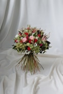 A vibrant bouquet of flowers set against a draped white fabric backdrop. The arrangement includes a variety of flowers in shades of red, pink, and white, interspersed with lush green foliage. The bouquet is tightly bound with long stems creating a rustic yet elegant appearance.