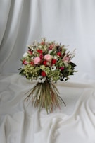 A vibrant bouquet of flowers set against a draped white fabric backdrop. The arrangement includes a variety of flowers in shades of red, pink, and white, interspersed with lush green foliage. The bouquet is tightly bound with long stems creating a rustic yet elegant appearance.