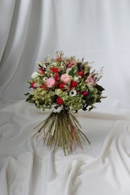 A vibrant bouquet of flowers set against a draped white fabric backdrop. The arrangement includes a variety of flowers in shades of red, pink, and white, interspersed with lush green foliage. The bouquet is tightly bound with long stems creating a rustic yet elegant appearance.