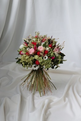 A vibrant bouquet of flowers set against a draped white fabric backdrop. The arrangement includes a variety of flowers in shades of red, pink, and white, interspersed with lush green foliage. The bouquet is tightly bound with long stems creating a rustic yet elegant appearance.