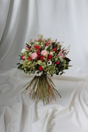 A vibrant bouquet of flowers set against a draped white fabric backdrop. The arrangement includes a variety of flowers in shades of red, pink, and white, interspersed with lush green foliage. The bouquet is tightly bound with long stems creating a rustic yet elegant appearance.
