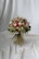 A vibrant bouquet of flowers set against a draped white fabric backdrop. The arrangement includes a variety of flowers in shades of red, pink, and white, interspersed with lush green foliage. The bouquet is tightly bound with long stems creating a rustic yet elegant appearance.