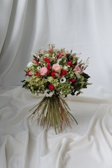 A vibrant bouquet of flowers set against a draped white fabric backdrop. The arrangement includes a variety of flowers in shades of red, pink, and white, interspersed with lush green foliage. The bouquet is tightly bound with long stems creating a rustic yet elegant appearance.
