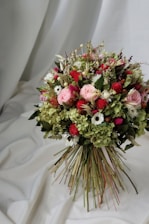 A beautiful bouquet composed of a variety of flowers, including roses and daisies, with a mix of soft pink, deep red, and white blooms. The arrangement is lush, with green foliage and textured leaves, creating a vibrant and natural look. The backdrop features a draped white fabric that complements the colors of the bouquet.
