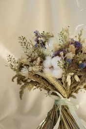 A bouquet of dried flowers and plants arranged with a light ribbon, featuring a variety of textures including fluffy cotton blooms, delicate wheat stalks, and small purple and white flowers against a softly blurred beige background.