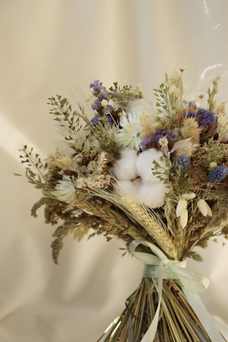 A bouquet of dried flowers and plants arranged with a light ribbon, featuring a variety of textures including fluffy cotton blooms, delicate wheat stalks, and small purple and white flowers against a softly blurred beige background.