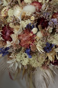An elaborate, close-up arrangement of dried flowers and foliage in various shades of red, purple, white, and beige. The bouquet features a mix of textures, including soft, fluffy elements, spiky and intricate dried flowers, and delicate leaves. The use of colors and textures creates a rich, organic aesthetic.