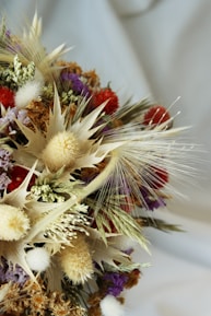 A close-up view of a dried flower bouquet featuring a variety of shapes and textures. The arrangement includes spiky, fluffy, and delicate elements in a mix of natural and vibrant colors. Feathery grass accents and a blend of beige, red, purple, and white tones create an intricate and visually interesting composition.
