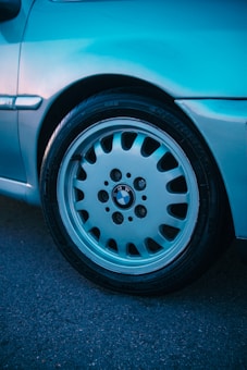 Close-up of a silver BMW car wheel with a prominent logo at the center, set against a smooth metallic body. The image captures the intricate design of the rim and the textured surface of the tire on a dark asphalt background.