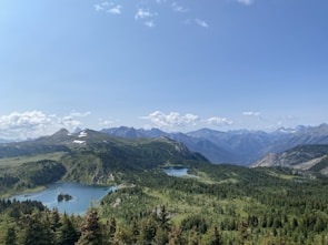 A vast landscape of lush green valleys and dense forests spread out under a clear blue sky. Snow-capped mountain peaks rise in the background, while a serene lake reflects the surrounding natural beauty.