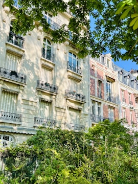 A row of elegant, multi-story residential buildings with classic architecture featuring ornate railings and large windows. Lush greenery and trees are in the foreground, with sunlight casting soft shadows on the facades.