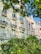 A row of elegant, multi-story residential buildings with classic architecture featuring ornate railings and large windows. Lush greenery and trees are in the foreground, with sunlight casting soft shadows on the facades.