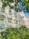 A row of elegant, multi-story residential buildings with classic architecture featuring ornate railings and large windows. Lush greenery and trees are in the foreground, with sunlight casting soft shadows on the facades.