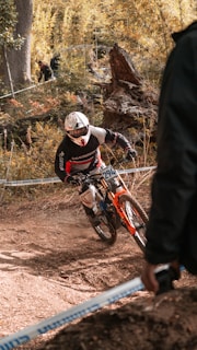 A mountain biker in full gear navigates a dirt trail through a wooded area. The biker rides a red and orange bike and appears to be participating in a competitive event, judged by the number on the bike and the presence of spectators in the background. The scene is set in autumn, with leaves scattered on the ground and fall foliage.