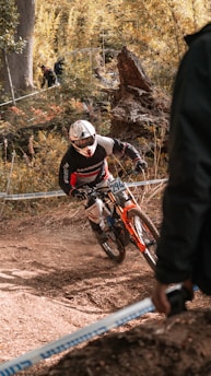 A mountain biker in full gear navigates a dirt trail through a wooded area. The biker rides a red and orange bike and appears to be participating in a competitive event, judged by the number on the bike and the presence of spectators in the background. The scene is set in autumn, with leaves scattered on the ground and fall foliage.