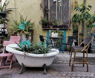 An outdoor scene featuring a vintage bathtub repurposed as a planter filled with greenery. Surrounding it are rustic elements like mismatched chairs and a small round table with white candles. The backdrop is a textured wall adorned with various plants and a decorative window frame. Earthy tones and eclectic decor create a serene, bohemian atmosphere.