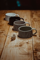 Four ceramic mugs of different colors are arranged in a line on a wooden table. The colors of the mugs include white, black, and two shades of grey. A small dried flower is placed next to the closest mug, adding a rustic element to the scene with soft lighting that casts gentle shadows.