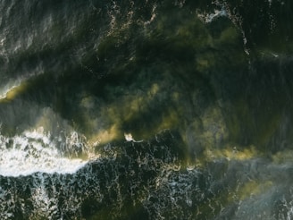 An aerial view captures turbulent ocean waves against a deep green backdrop. Swirls of foam and lighter patches indicate areas of disturbance, suggesting strong currents or wind activity.
