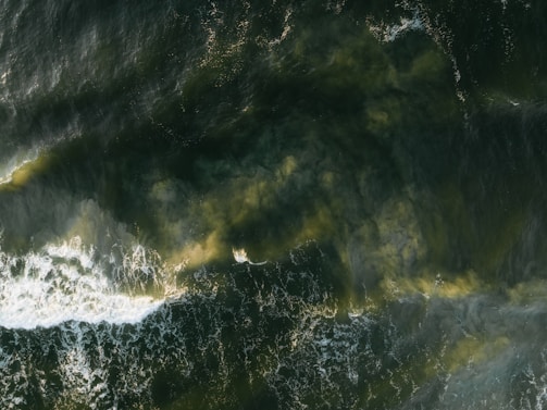 An aerial view captures turbulent ocean waves against a deep green backdrop. Swirls of foam and lighter patches indicate areas of disturbance, suggesting strong currents or wind activity.
