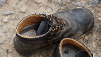A pair of worn, black work boots with yellow and black laces are resting on a rough, gravelly surface. The insides of the boots show the yellow lining and the dotted insoles.