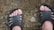 A pair of feet wearing black slip-on sandals stands on a rocky and uneven ground surface. The ground consists of dirt and textured stones, giving an earthy and rugged appearance.