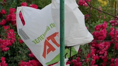 A white plastic bag with printed green and red text hangs on a green metal pole. The background features vibrant pink flowers and green foliage.