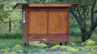 A small wooden shed with a slanted roof is surrounded by lush greenery and trees. The structure is elevated on short stilts and has a weathered, natural finish. A window is visible on the side, and yellow wildflowers are in the foreground.