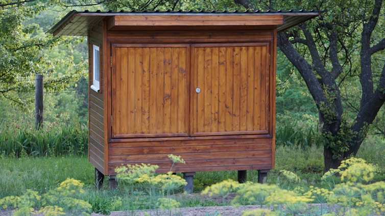A small wooden shed with a slanted roof is surrounded by lush greenery and trees. The structure is elevated on short stilts and has a weathered, natural finish. A window is visible on the side, and yellow wildflowers are in the foreground.