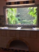 A kitchen sink area adorned with intricate ceramic tiles featuring blue and yellow patterns. The window above the sink offers a view of lush green plants and a stone wall covered in ivy, creating a seamless blend between indoors and outdoors.