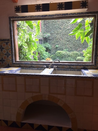 A kitchen sink area adorned with intricate ceramic tiles featuring blue and yellow patterns. The window above the sink offers a view of lush green plants and a stone wall covered in ivy, creating a seamless blend between indoors and outdoors.