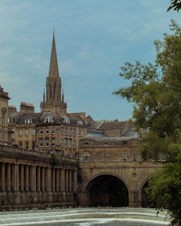 A picturesque scene featuring a historical bridge with an arched design spanning a calm river, complemented by cascading water in the foreground. Elegant stone buildings with numerous windows line one side of the river, while a prominent spire rises into the sky in the background. Lush green trees frame the right side of the image, adding a touch of nature to the urban setting.